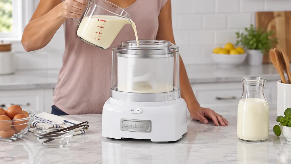 A hand pouring chilled vanilla base through the pour spout of a running Cuisinart ice cream maker, the paddle already turning inside the frozen bowl.