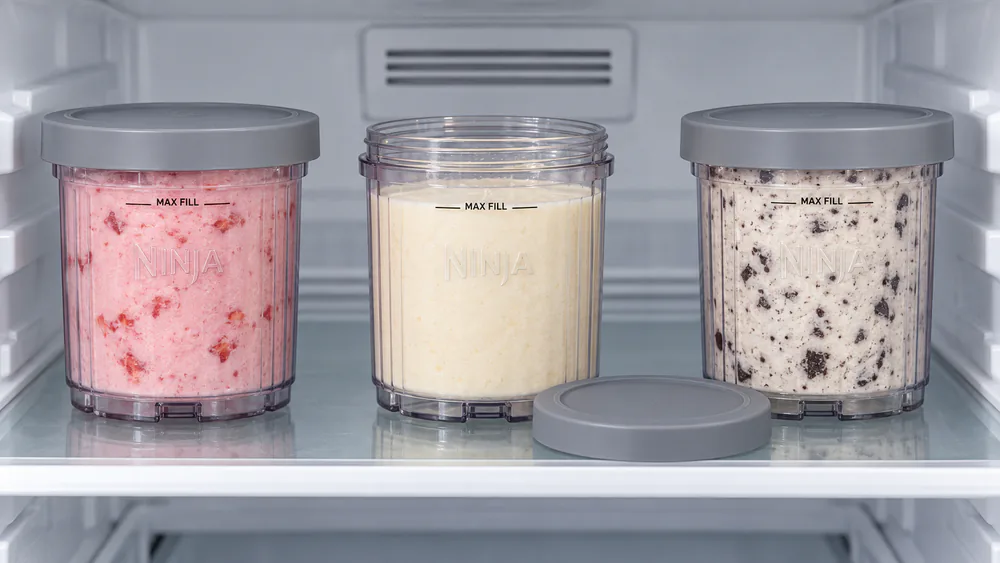 A Ninja Creami Swirl pint with its lid off, level on a freezer shelf beside two more filled pints with storage lids on.