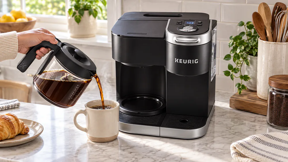 A hand pouring from the Keurig K-Duo Gen 1 glass carafe into a mug, a small dribble of coffee running down the carafe spout and side.