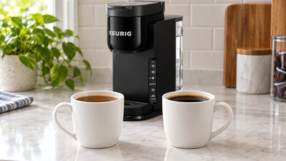 Two cups of coffee on a kitchen counter, one brewed regular and one with Strong Brew, the Strong Brew cup visibly darker, with the compact K-Express brewer in the background.