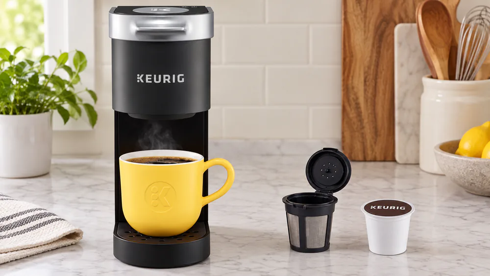 A mug of coffee next to a Keurig K-Mini, with a reusable My K-Cup filter and a K-Cup pod.
