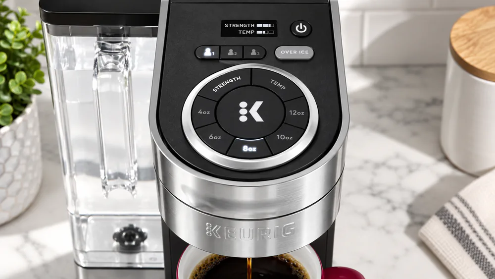 Close-up of the Keurig K-Supreme Plus control panel showing the digital display, the size, strength, and temperature buttons, and the lit brew cycle, on a kitchen counter.