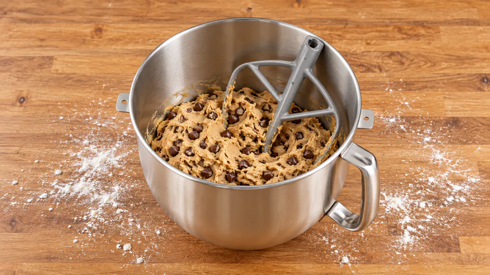 A large double batch of chocolate chip cookie dough in the 7-quart stainless bowl with the coated flat beater, flour dusted on the counter beside it