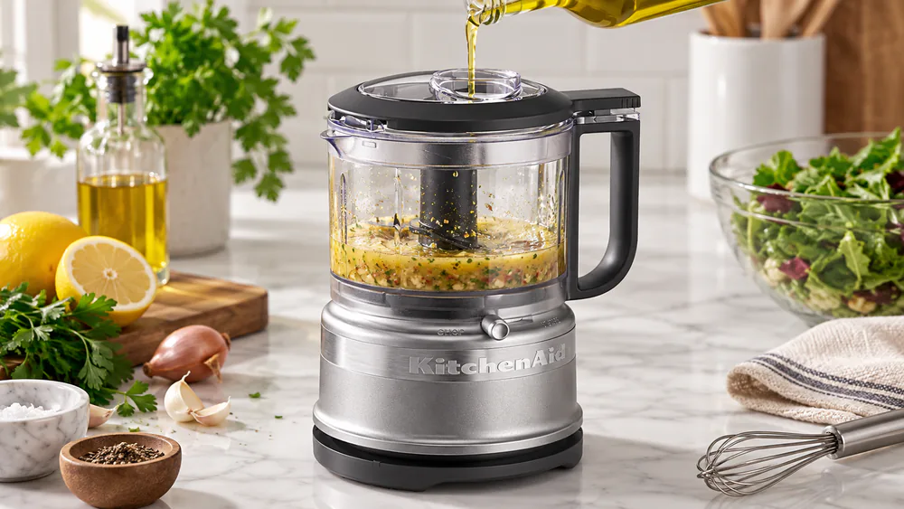 Oil streaming through the drizzle basin of a KitchenAid food chopper lid into an emulsifying vinaigrette below.