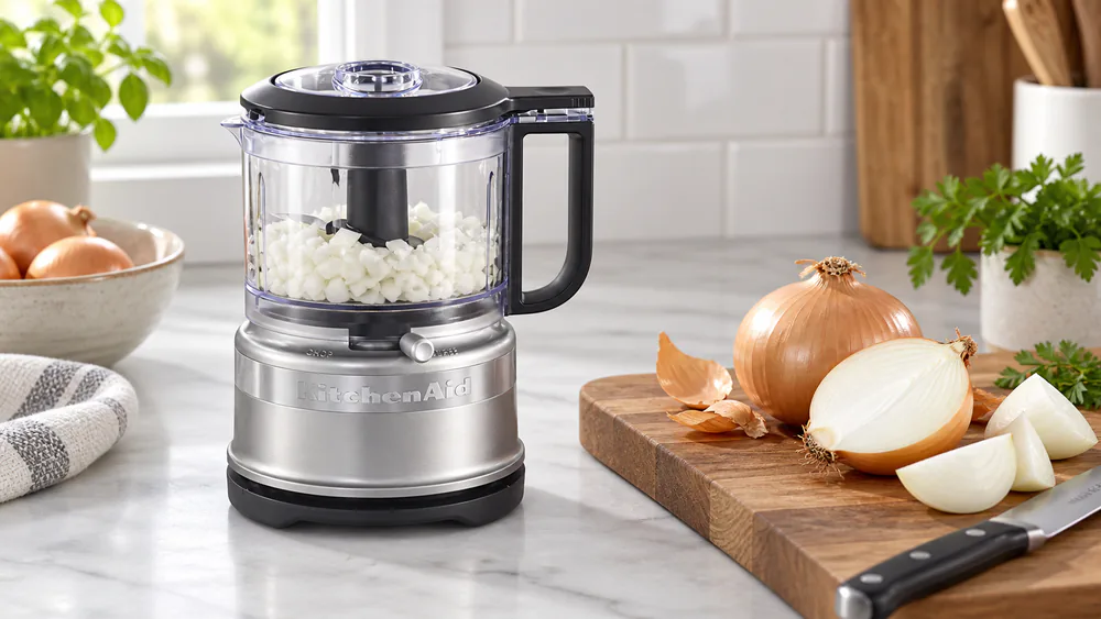 A KitchenAid 3.5 cup food chopper bowl filled with evenly chopped white onion next to a cutting board with whole onions.