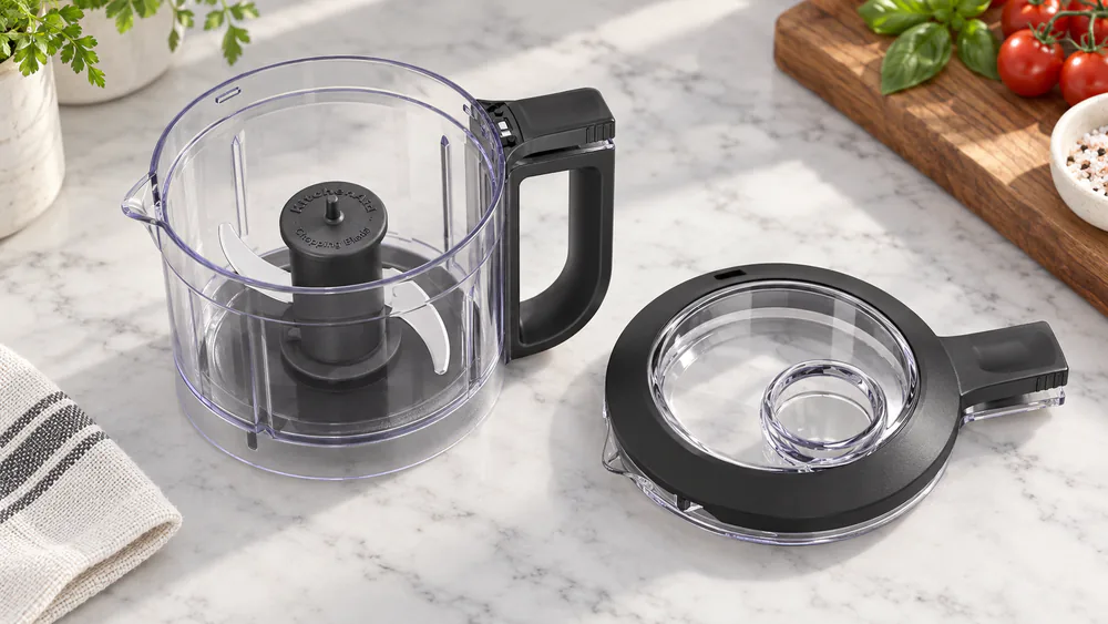 The three parts of a KitchenAid 3.5 cup food chopper laid out on a counter: the work bowl, the stainless steel blade, and the lid with drizzle basin.