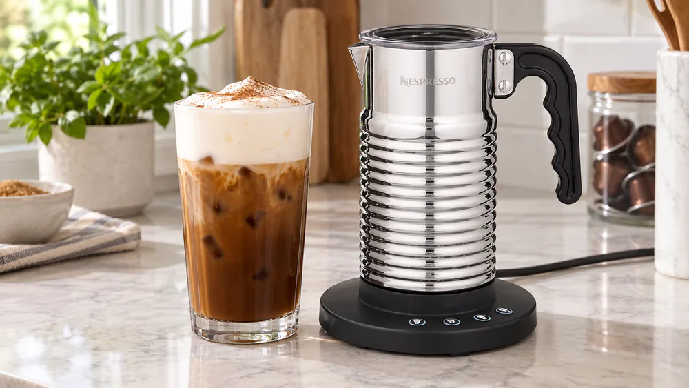 A tall glass of iced coffee topped with a thick layer of cold foam made in the Nespresso Aeroccino 4, the frother jug beside it on the counter.
