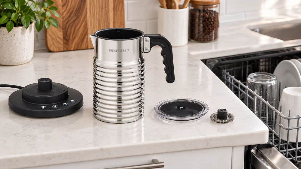 The Nespresso Aeroccino 4 frothing jug, lid, and magnetic whisk separated on a kitchen counter beside an open dishwasher rack.