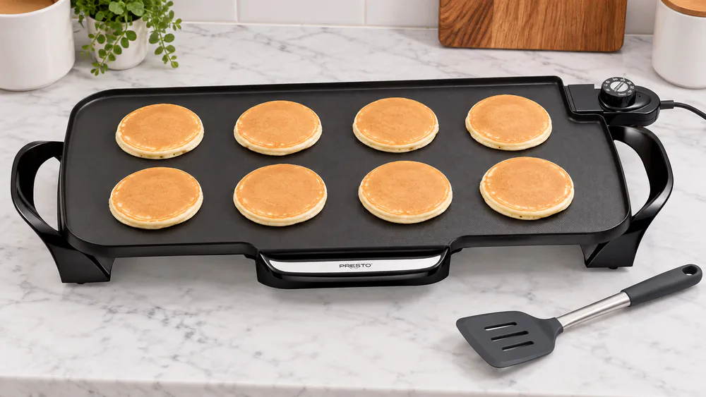 Eight pancakes cooking on the Presto 22-inch ceramic griddle with a silicone spatula resting at the edge