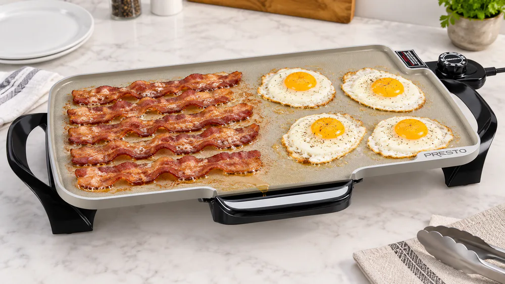 Strips of bacon and fried eggs cooking side by side on a Presto 07062 griddle, grease running toward the drip tray