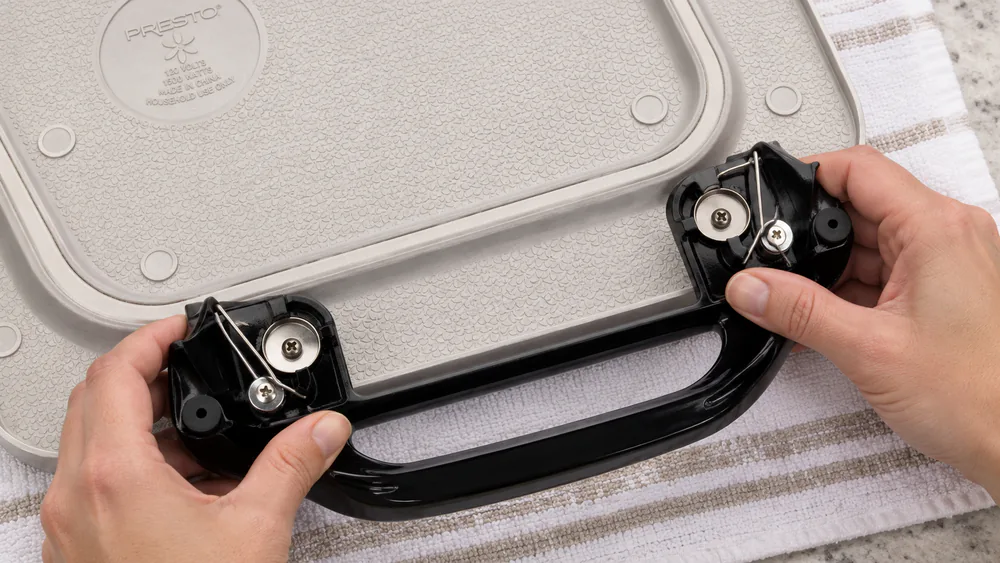 Close-up of a Presto 07062 griddle with one handle detached, showing the latch clip on the cast aluminum base