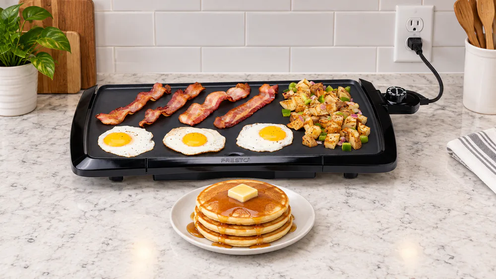 Presto 07030 electric griddle on a family kitchen counter with a short cord plugged into a nearby outlet and a stack of pancakes on a plate