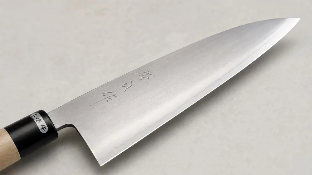 Close-up of the Toyama 210mm gyuto convex grind and kasumi cladding under kitchen light, showing the thin edge and satin flat.