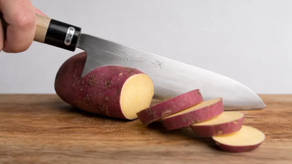 The Toyama 210mm gyuto mid-cut through a dense sweet potato on a wooden board, clean separated slices falling away.
