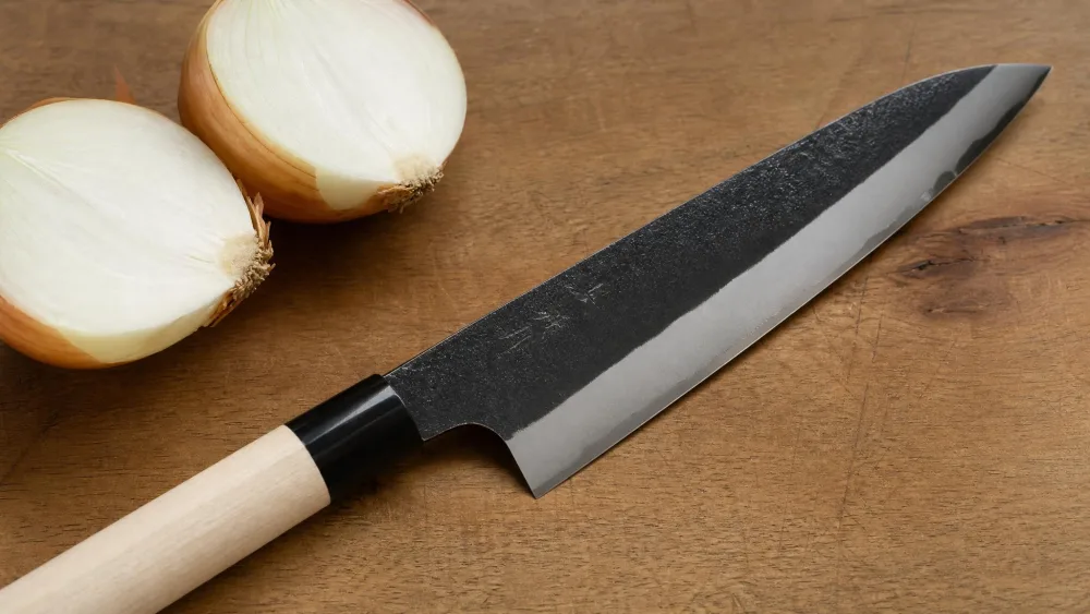 A Wakui Blue #2 kurouchi nashiji gyuto lying on a wooden cutting board beside halved onions, the dark forged finish above a bright polished bevel.