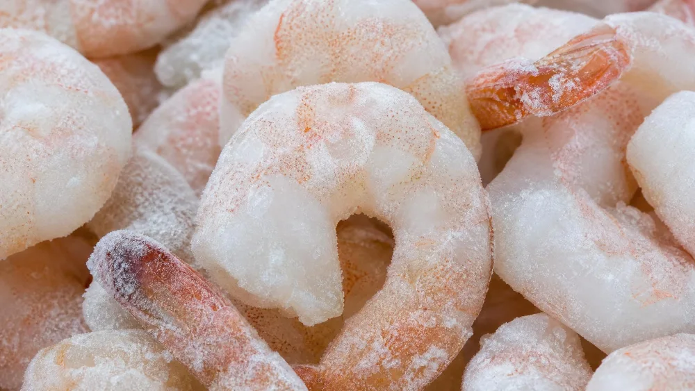 A macro shot of individually quick-frozen shrimp under a thin white ice glaze, frosty coating visible across the pieces.