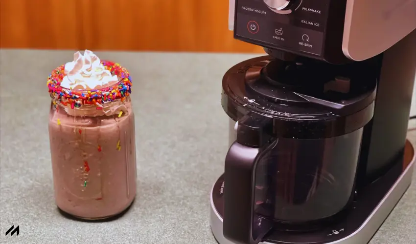 A decorated mason jar filled with a creamy milkshake topped with whipped cream and sprinkles sits next to a dessert machine.