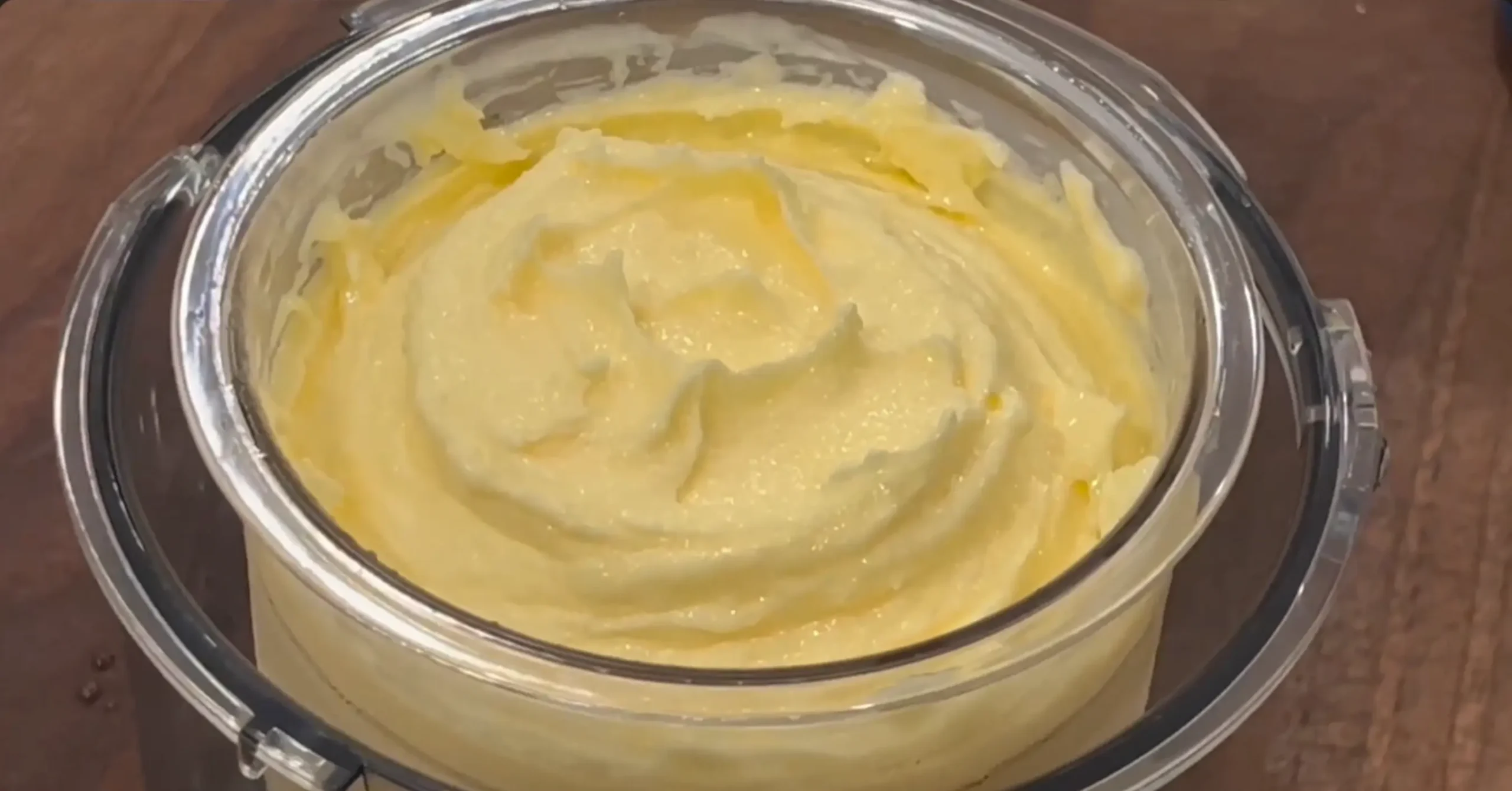 A close-up view of creamy yellow butter or Dole whipped frosting in a clear mixing bowl, resting on a wooden surface.