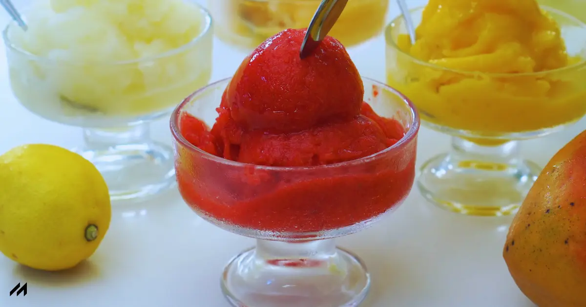 A vibrant red sorbet in a glass dish, surrounded by tropical fruit sorbets and a lemon on a white background.