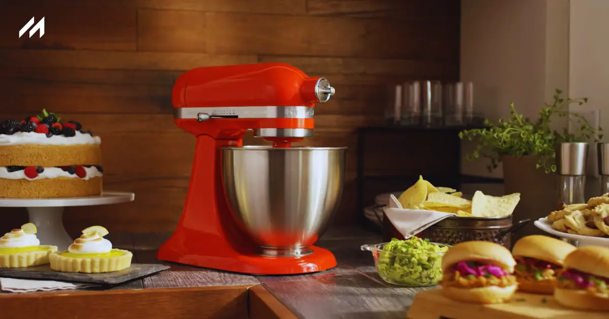 A vibrant red stand mixer sits on a wooden countertop, surrounded by desserts, guacamole, and tacos, in a cozy kitchen setting.