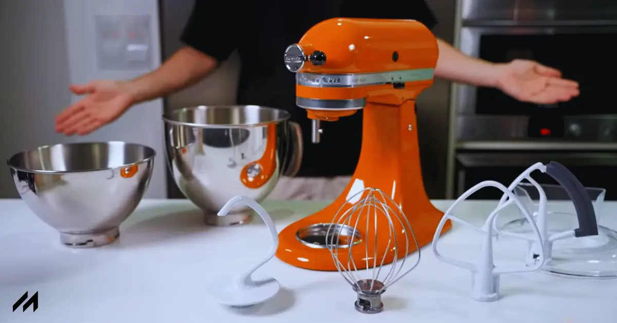 A vibrant orange Kitchenaid stand mixer on a countertop with two stainless steel mixing bowls and various attachments displayed around it.