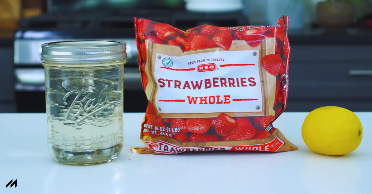 A mason jar with water, a bag of frozen whole strawberries, and a yellow lemon on a kitchen countertop.