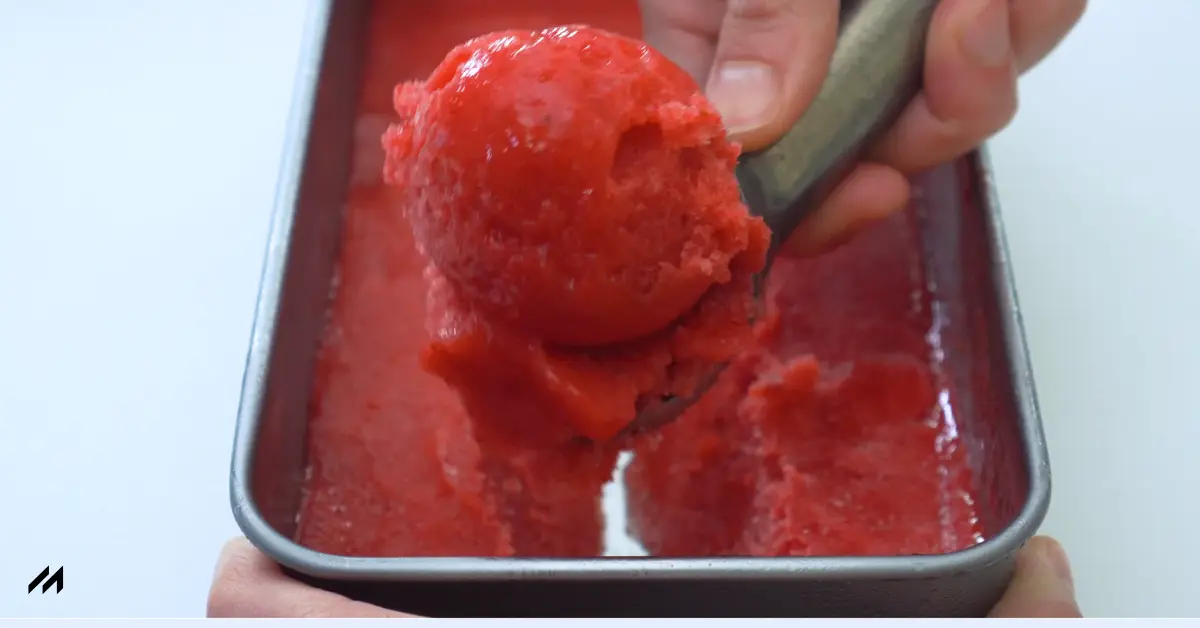 A hand scooping a vibrant red sorbet from a metal container, showcasing its smooth texture and rich color.