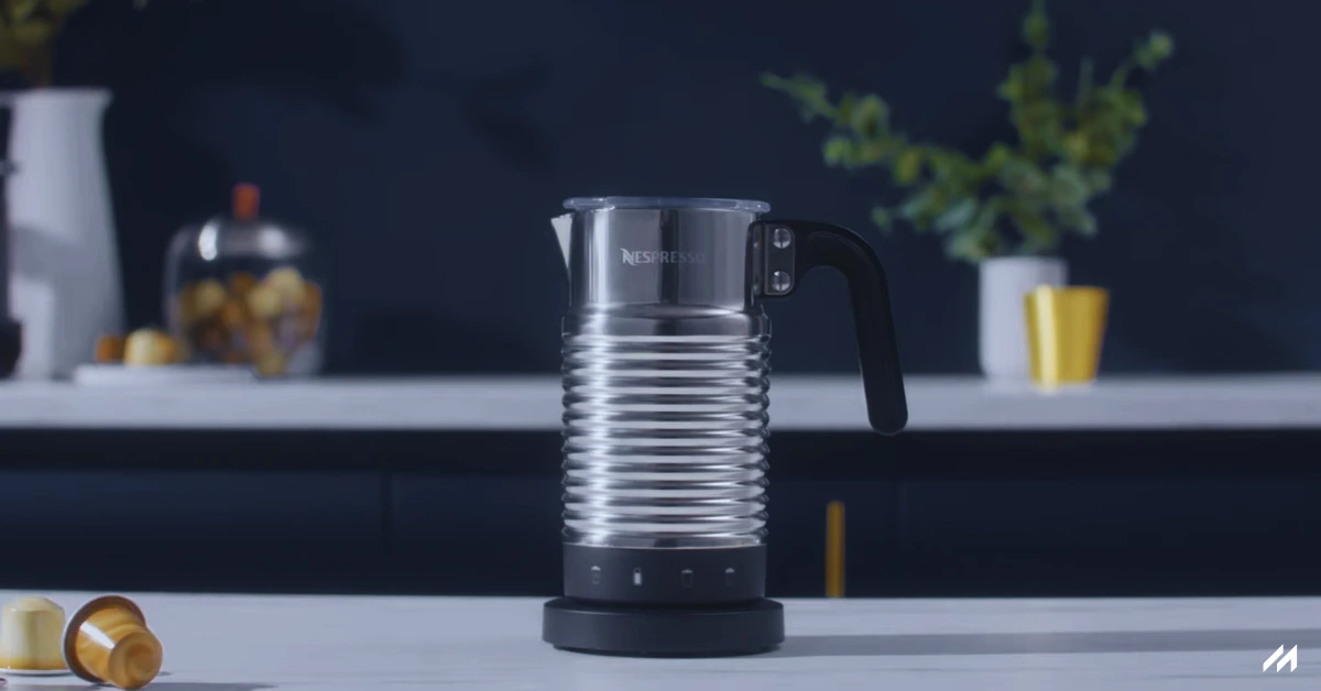 The image shows a Nespresso Aeroccino 4 milk frother with a sleek stainless steel body and a black ergonomic handle, placed on a modern kitchen countertop. The frother is positioned against a deep blue background with a few plants and colorful objects on the table, adding a stylish touch to the scene.