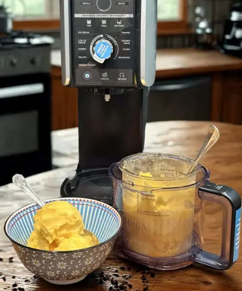 Preparing Ingredients for Ninja Creami A Ninja ice cream maker with a bowl of yellow ice cream beside it, sitting on a wooden table in a cozy kitchen.