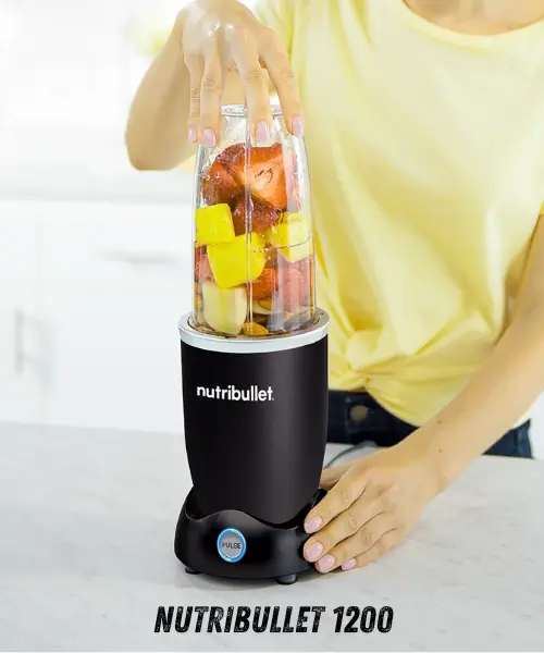 A person in a yellow shirt prepares a NutriBullet 1200 blender, filled with fruits and nuts, for blending on a kitchen countertop.