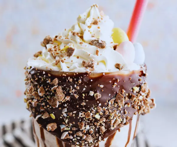A decadent ice cream sundae topped with whipped cream, chocolate drizzle, crushed nuts, and colorful candy, served with a straw.