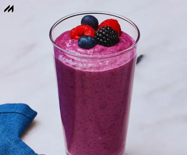 A tall glass of vibrant purple smoothie topped with raspberries, blueberries, and blackberries, set on a marble surface with a blue napkin.