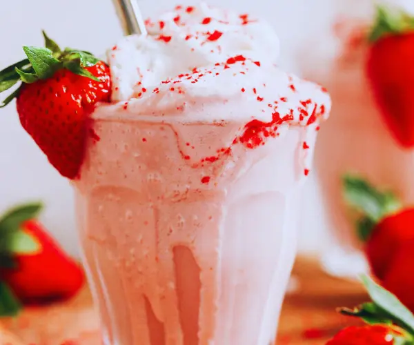 A glass of creamy strawberry milkshake topped with whipped cream, strawberry bits, and a fresh strawberry garnish.