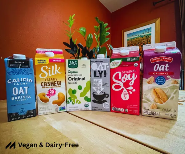 A lineup of six dairy-free milk cartons: oat, soy, and cashew varieties, with a plant and artwork in the background.