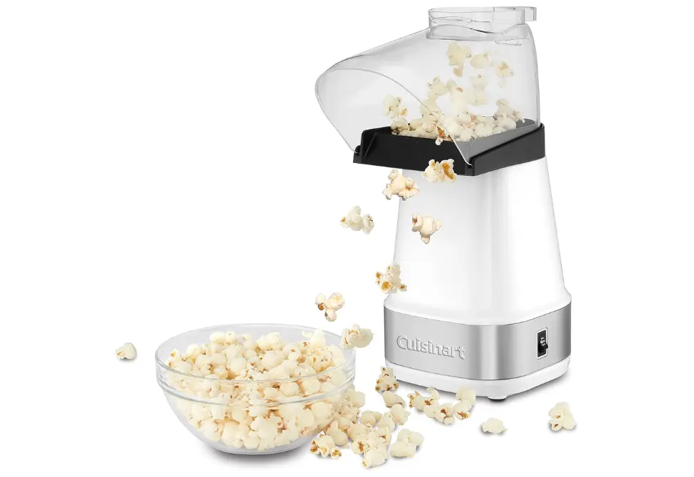 White Cuisinart air popper machine ejecting freshly popped popcorn into a clear glass bowl, with additional popcorn pieces scattered around on the surface