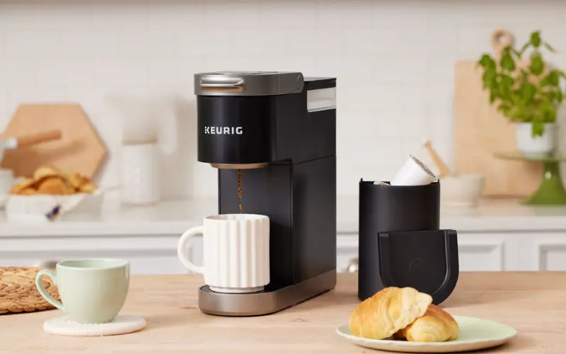 Keurig K-Mini Plus Coffee Maker brewing coffee into a white mug with croissants on a kitchen countertop.