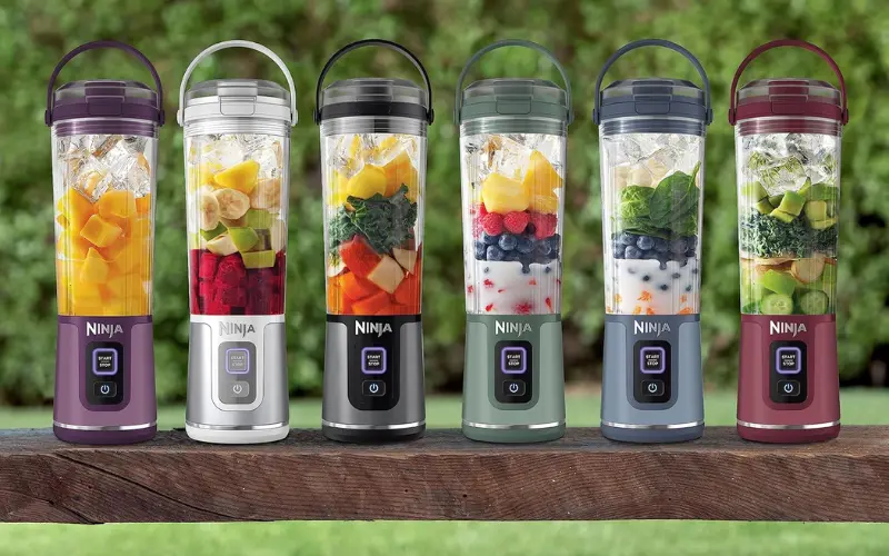 Six Ninja Blast portable blenders in different colors lined up on a wooden bench, each filled with fruit, greens, ice, or smoothie ingredients.