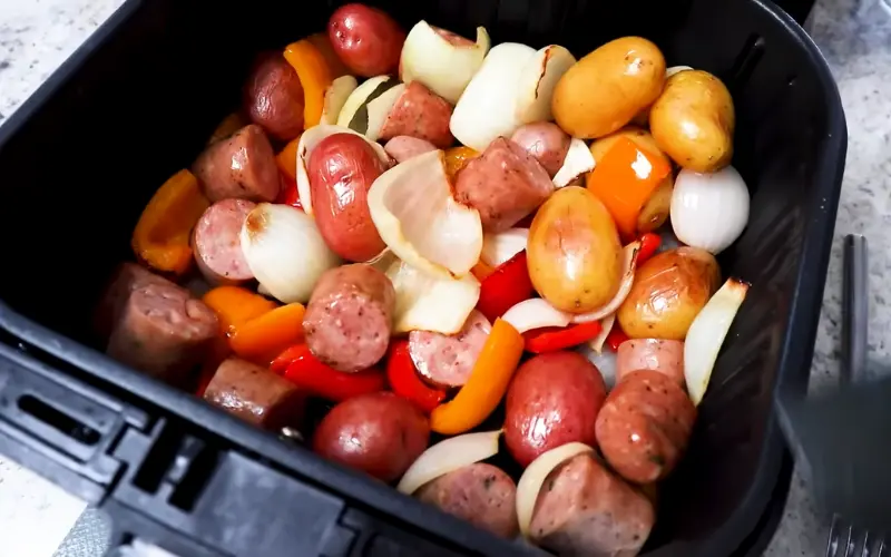 Air Fryer Sausage, Potatoes, and Onions Recipe Air fryer basket filled with sliced sausage, baby potatoes, colorful bell peppers, and onions ready to cook